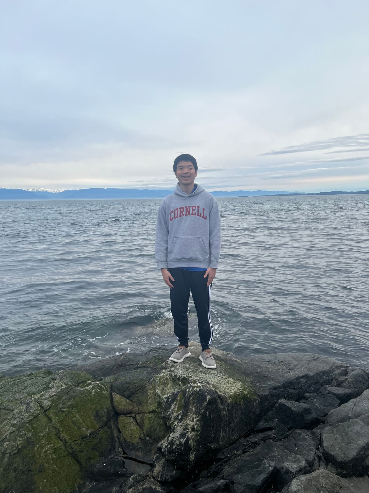 Shao standing on rocks at the water's edge in Alaska, mountains across the inlet behind him.