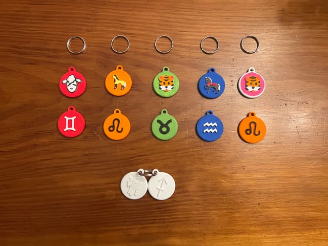 Twelve multi-colour 3D-printed zodiac keychains laid out in rows on a wooden table.